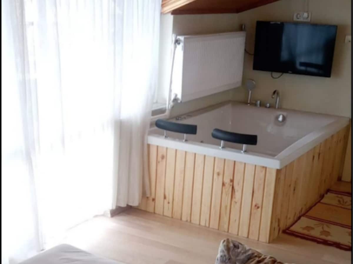 An inviting corner of a room features a modern bathtub with a wooden surround, accompanied by two sleek black chairs. Natural light filters through sheer white curtains, illuminating the space. A flat-screen TV is mounted on the wall nearby, enhancing the comfort of the area.