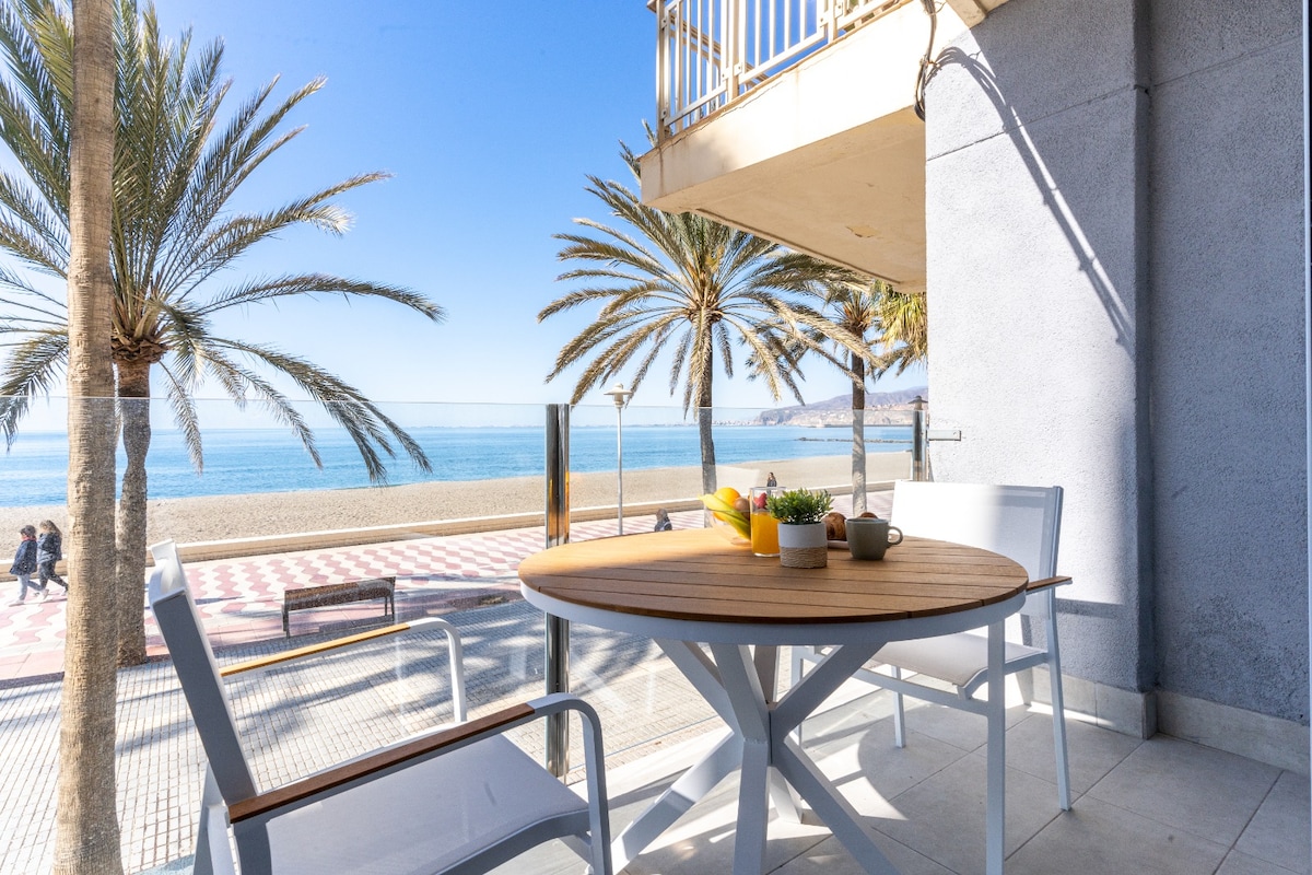 A terrace area features a round wooden table surrounded by four white chairs, offering a view of palm trees along the beach. The calm Mediterranean Sea is visible in the background, with a clear blue sky above. A small decorative plant and two cups are placed on the table.