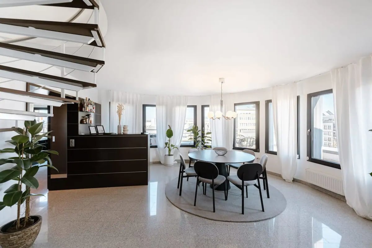 A stylish dining area features a round table with four black chairs, set on a soft circular rug. Large windows adorned with sheer white curtains frame the view, while potted plants add a touch of greenery to the space. A sleek bar area blends seamlessly with the elegant decor.