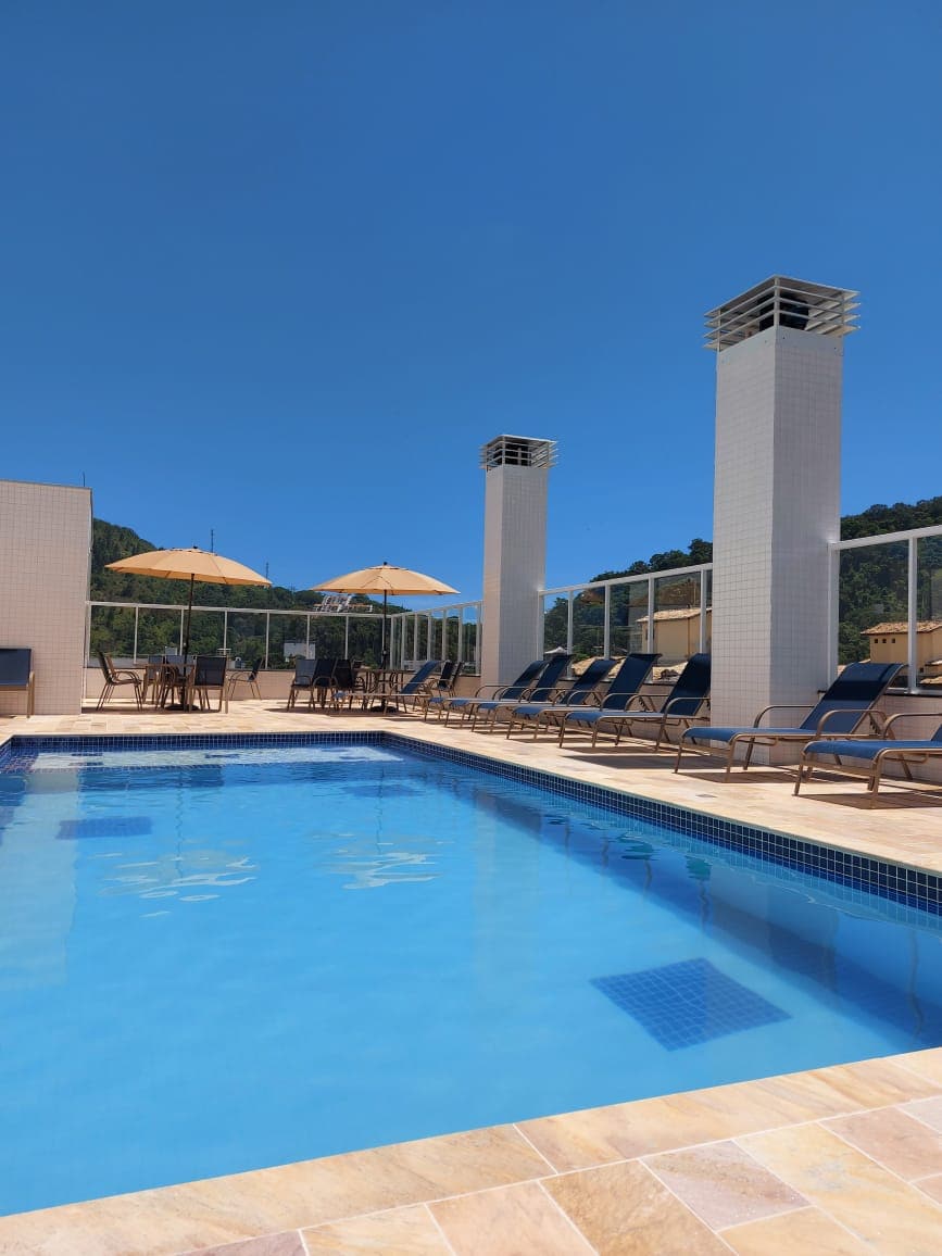 A rooftop pool area features a clear blue pool surrounded by lounge chairs and umbrellas. Tall white columns rise alongside the pool, and a scenic view of the mountains is visible in the background, adding to the relaxing atmosphere.