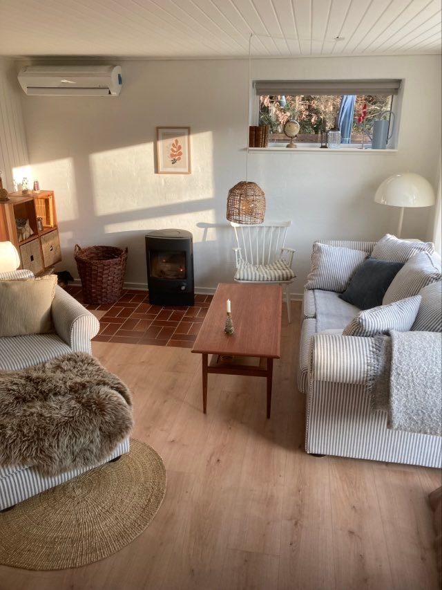 A bright living area is highlighted by a cozy seating arrangement, including two striped sofas and a wooden coffee table. A small wood-burning stove provides warmth, while natural light filters in through a window, creating a relaxing ambiance.