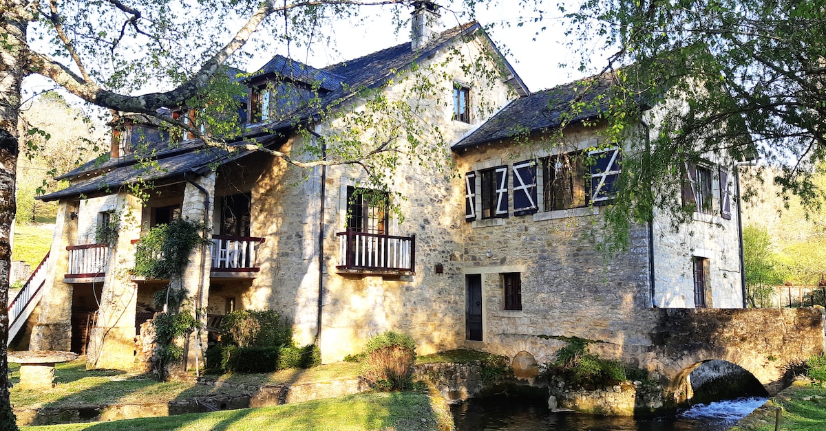 A charming stone house is set against a natural backdrop, featuring several large windows and balconies. The property is bordered by a peaceful river and includes an old mill structure, enhancing the serene atmosphere. Lush greenery surrounds the outdoor area, highlighting the tranquil environment.