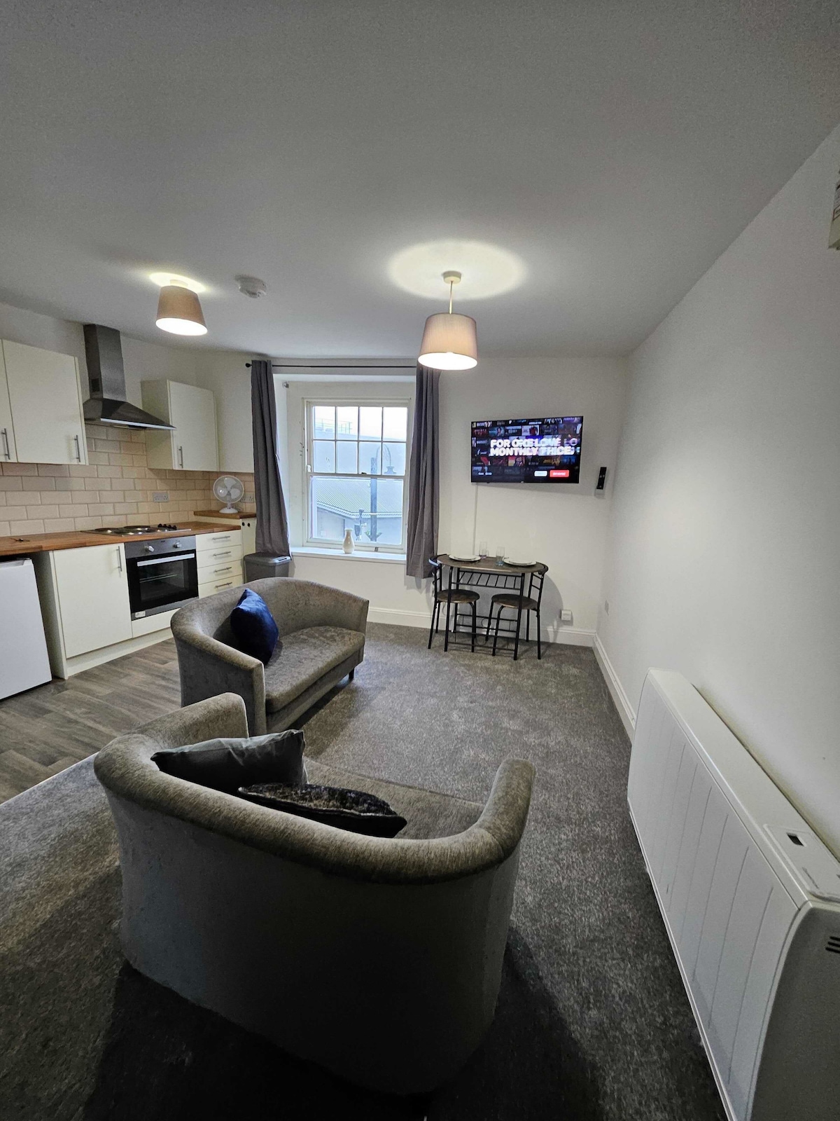 A combined kitchen and living area features two comfortable armchairs facing a flat-screen TV mounted on the wall. A small dining table with two chairs is positioned near a window, allowing natural light to fill the space.