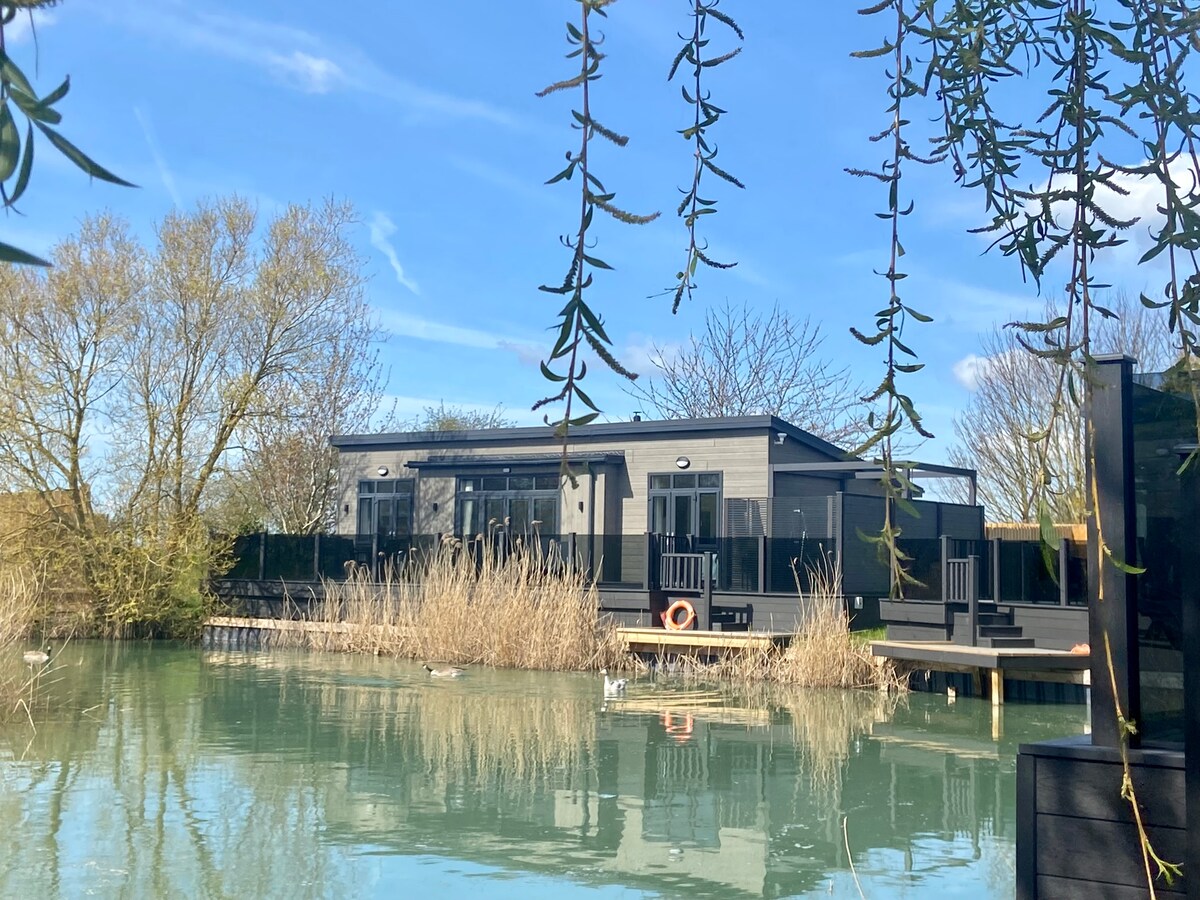 A stylish lodge is set against a serene lake, framed by gently swaying foliage. The building features large bi-fold doors that open towards a spacious deck, reflecting the tranquil surroundings and inviting moments of relaxation by the water.
