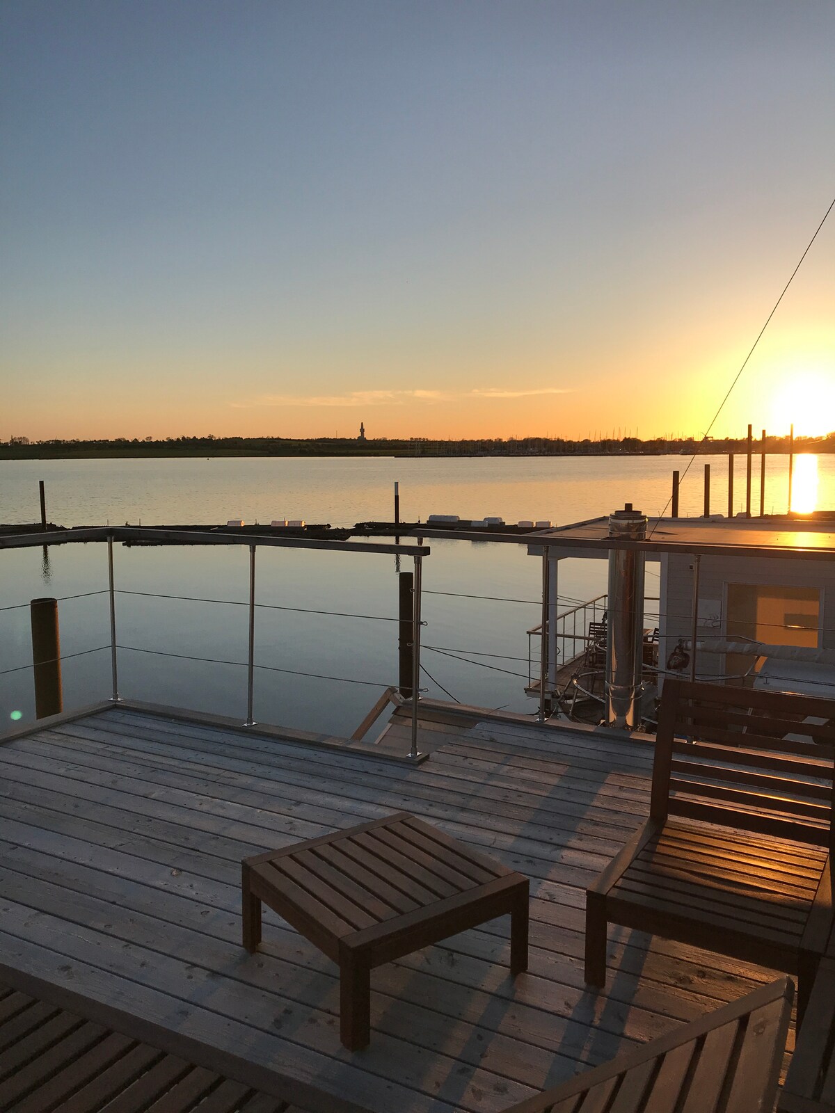 A furnished terrace is seen overlooking the water, where a wooden table and chairs are positioned for relaxation. The sunset casts a warm glow over the calm surface of the water, creating a serene backdrop for peaceful moments.
