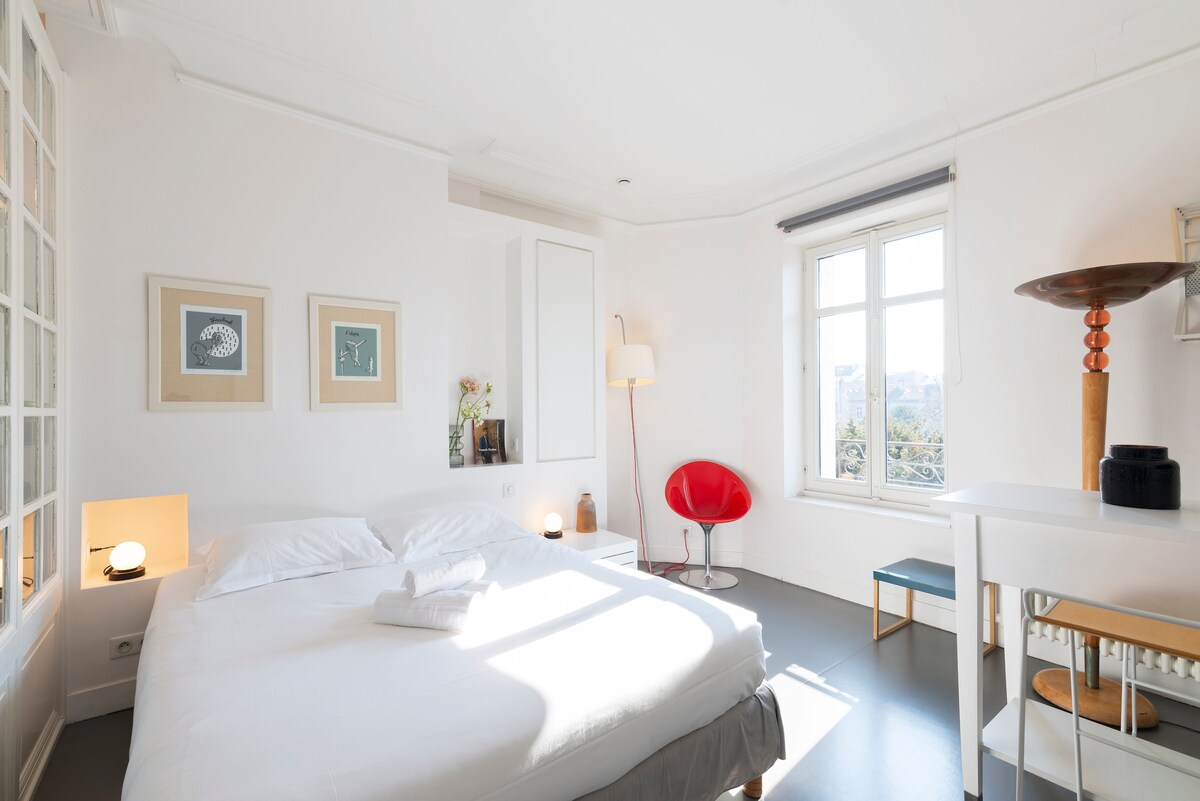 A well-lit bedroom features a spacious bed with white linens and towels neatly placed on top. Two framed artworks adorn the light-colored walls. A modern, red accent chair and a unique side table are positioned near a tall window, which allows natural light to fill the room.