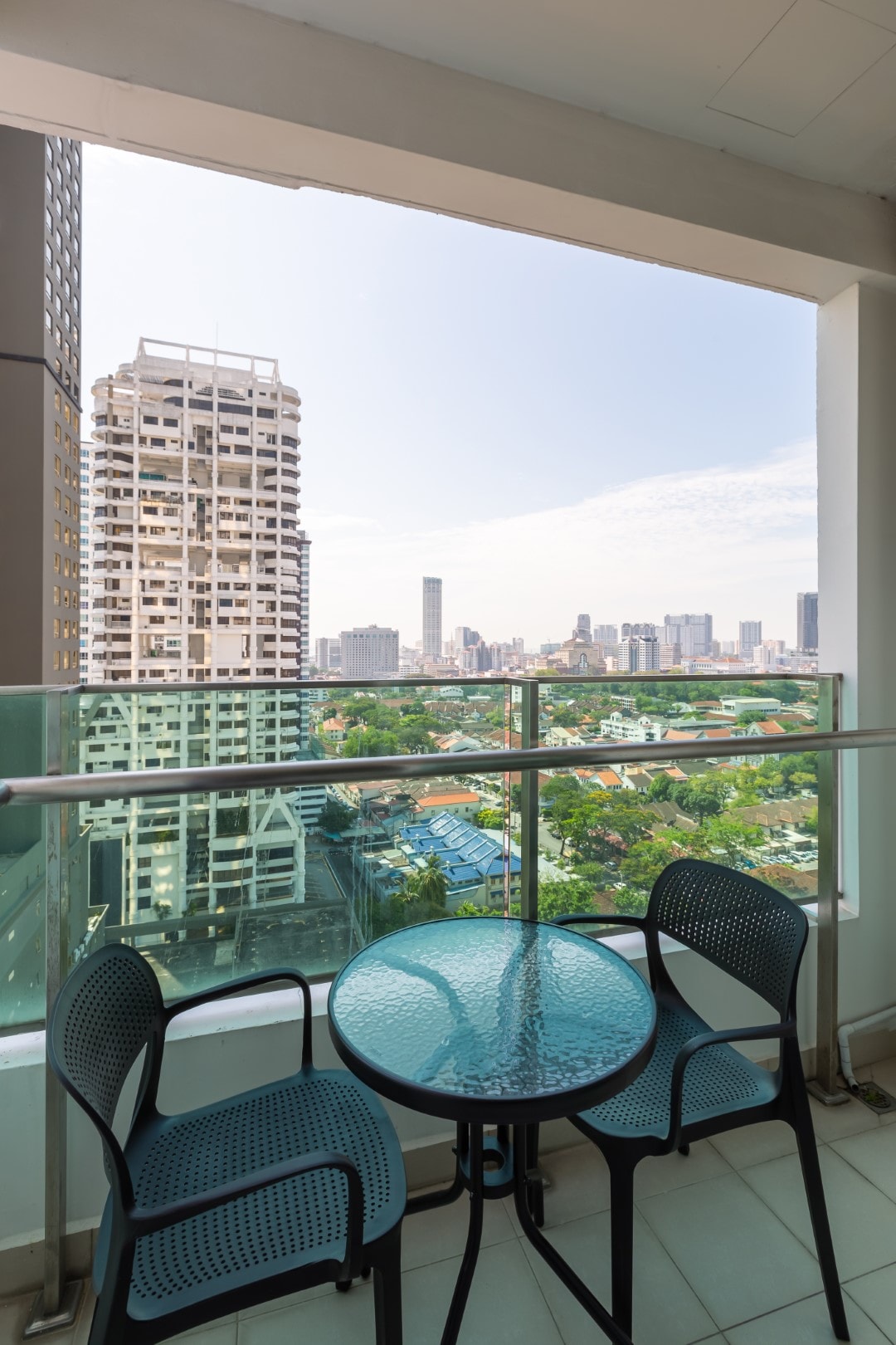 A small balcony features a round glass table surrounded by two black chairs. The view encompasses a vibrant cityscape below, with buildings and greenery visible in the distance, revealing a blend of urban and natural elements.