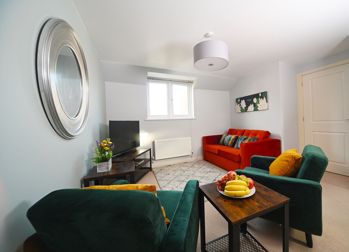An open-plan living area features a bright, modern space with a large circular mirror on the wall. A red sofa and a green armchair frame a small table adorned with fresh fruit. Natural light enters through a window, illuminating the contemporary decor.