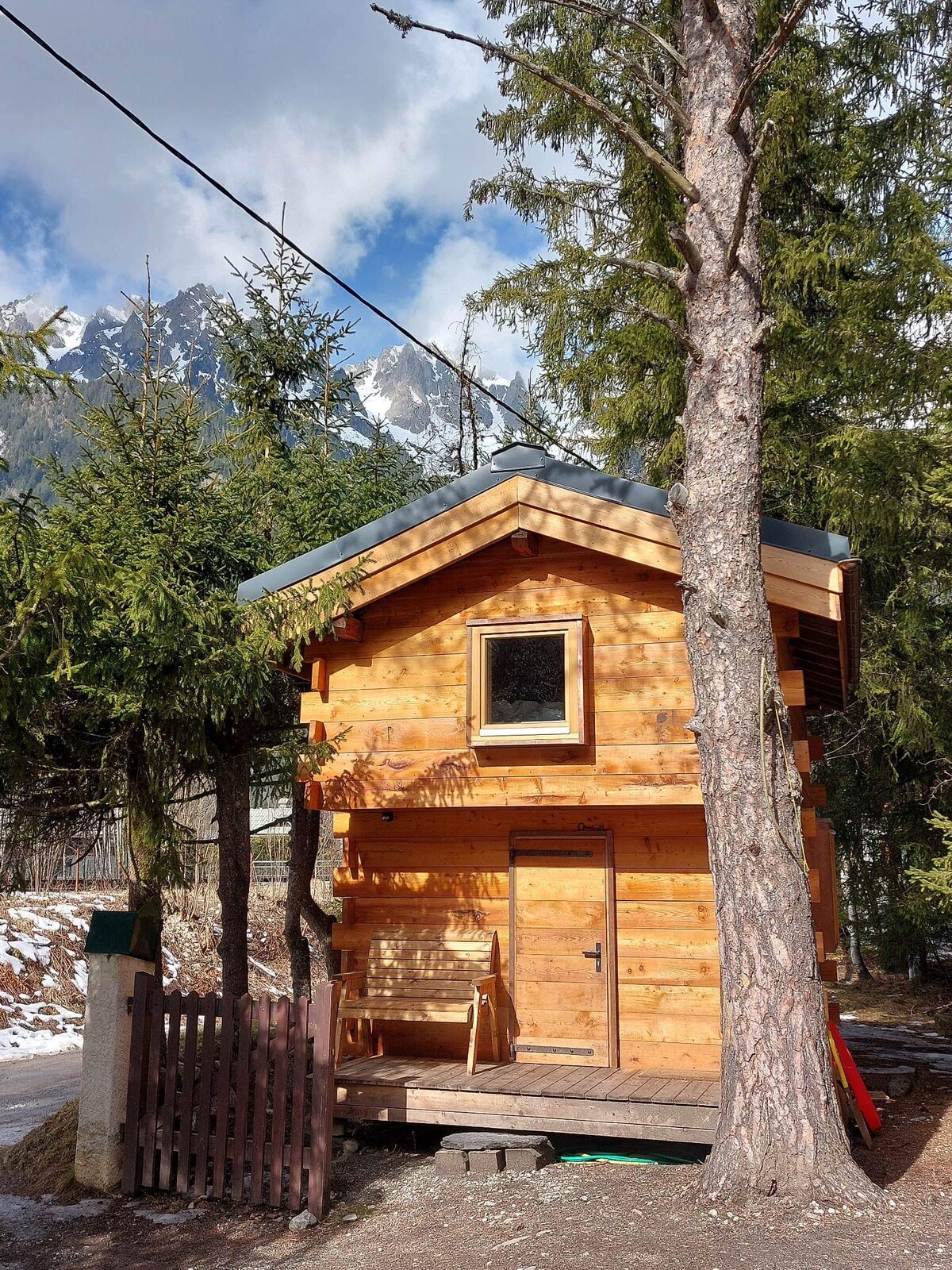A charming wooden cabin features a sloped roof and a large window on the upper level. Surrounded by tall trees, the cabin is nestled in a natural setting, with snow-capped mountains visible in the background. A small porch area is seen at the front.
