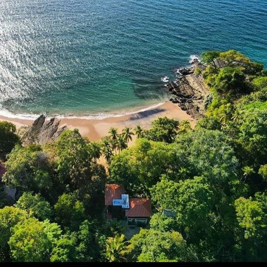Aerial view reveals a secluded sandy beach bordered by lush green foliage and trees. Gentle waves lap at the shore, with rocky outcrops scattered along the coastline. A small structure with a reddish roof can be seen amidst the vibrant vegetation, enhancing the natural beauty of the scene.