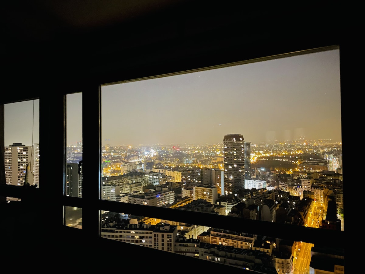 The view from the apartment showcases a panoramic cityscape illuminated at night, featuring twinkling lights and the distant outline of skyscrapers. A large window frames this vibrant scene, offering insight into the bustling urban environment below.
