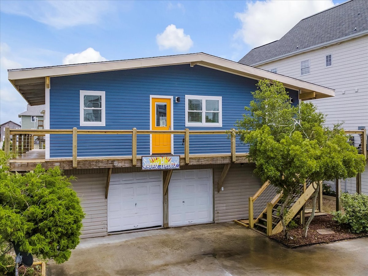 A bright blue house features a welcoming orange door, elevated above ground level. A wooden deck extends from the front, surrounded by greenery. Stairs lead to the entrance, and a banner is displayed prominently. The ground area includes a parking space beneath.