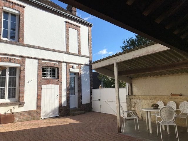 An outdoor courtyard is shown, featuring a section with a covered area and a table surrounded by four white chairs. The brick exterior of the house is visible, complemented by a clear blue sky overhead.