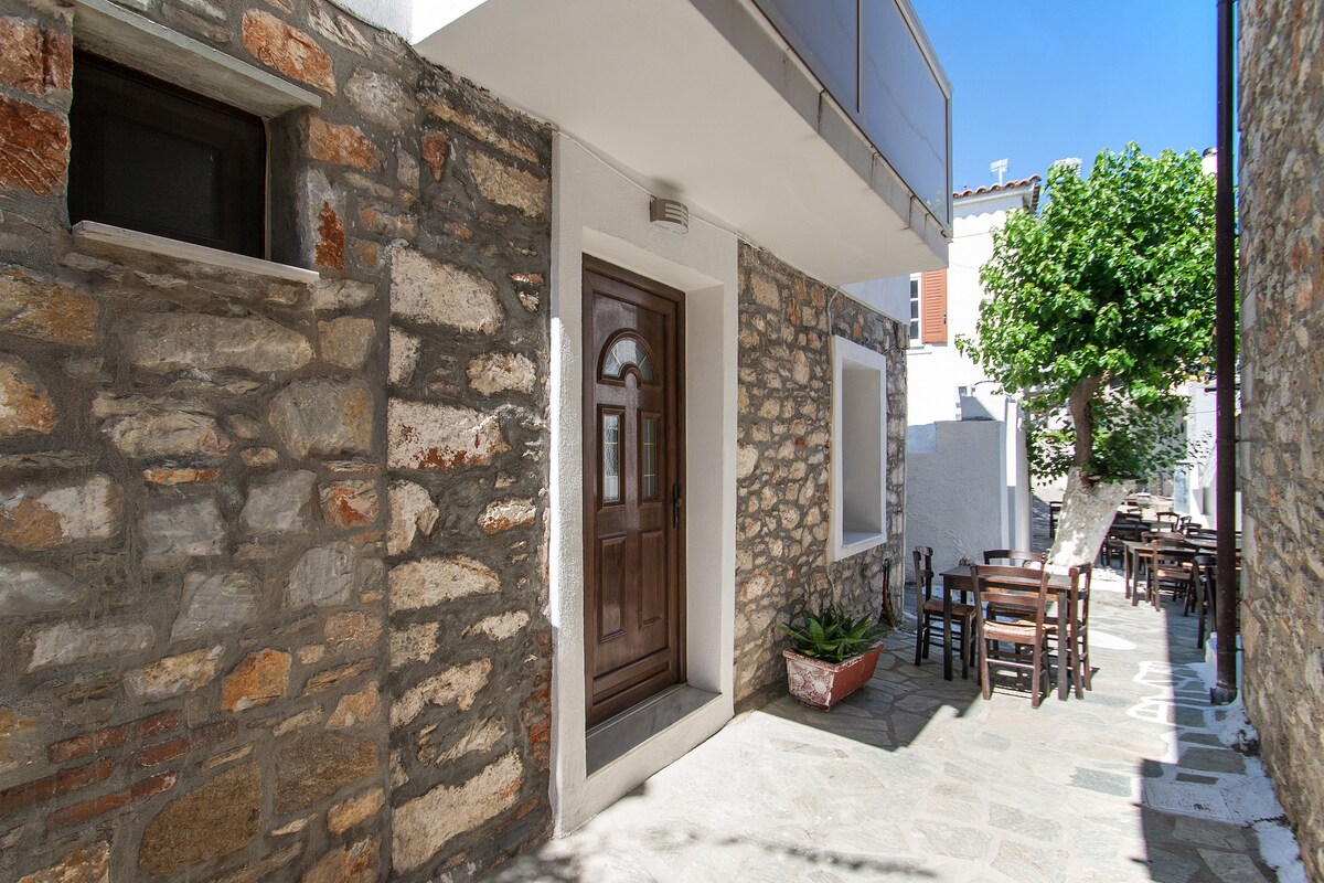 A charming stone pathway leads to the entrance of the apartment, framed by rustic walls and greenery. Outdoor seating is arranged at wooden tables along the pathway, offering a community feel. Sunlight brightens the scene, highlighting the texture of the stone and the wooden door.