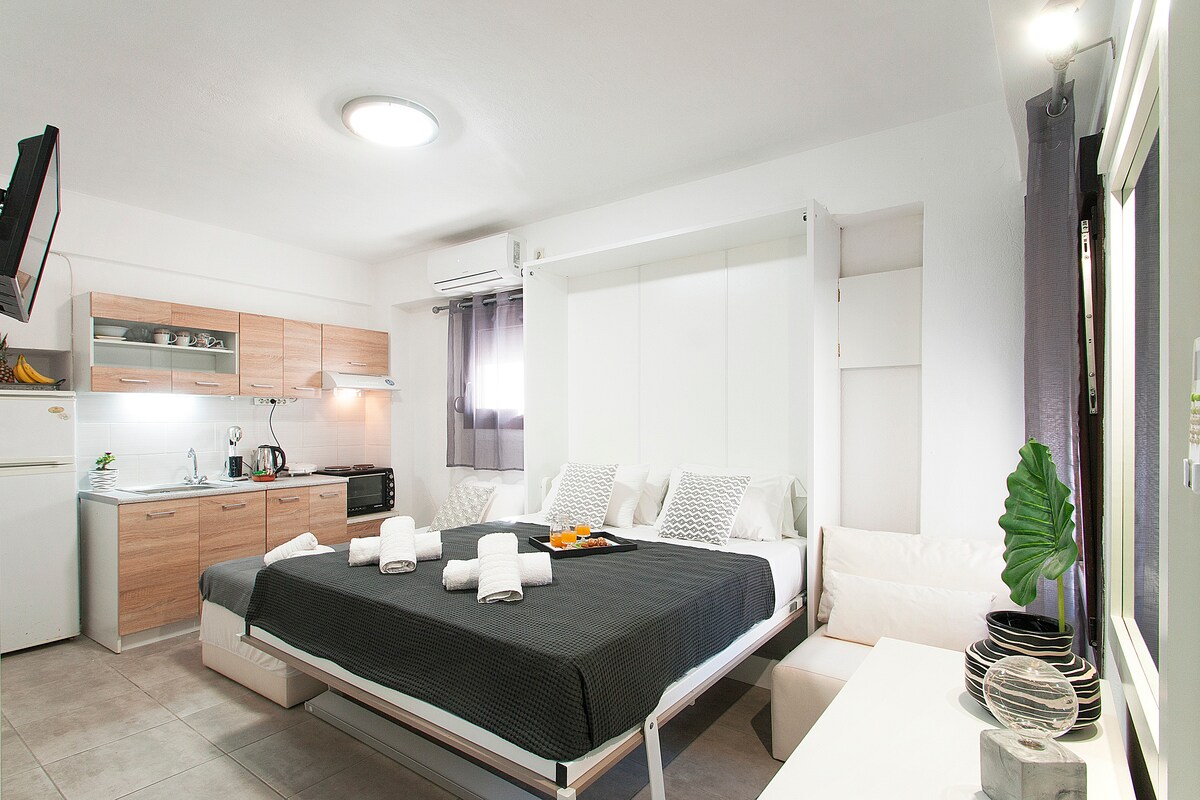 A modern studio apartment features a spacious bed equipped with a light grey bedspread. A kitchenette with wooden cabinetry is visible in the background, alongside a dining area that includes a table and white seating. Natural light flows through the window, enhancing the bright interior.
