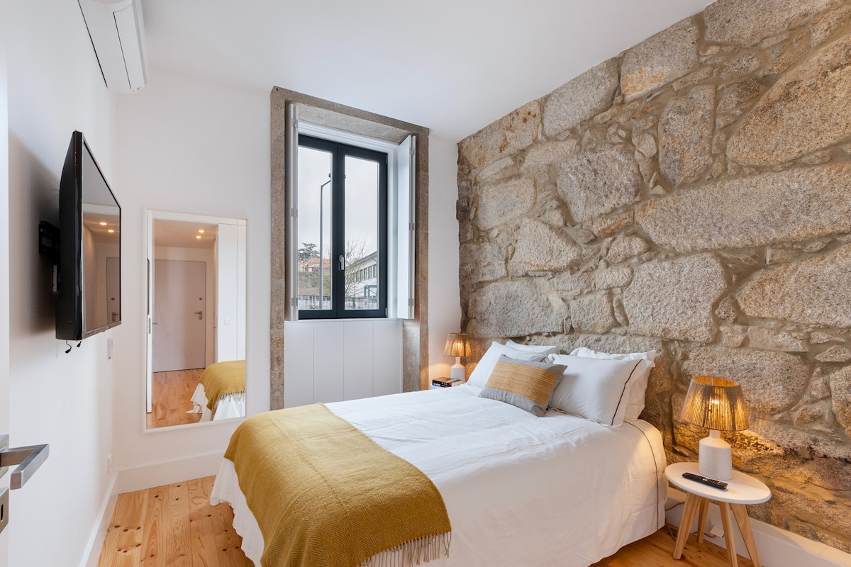 A cozy bedroom displays a comfortable bed with light bedding and a mustard-colored throw. Exposed stone walls lend texture, and natural light filters through the large window. A modern TV is mounted on the wall, while bedside tables with lamps enhance the space.