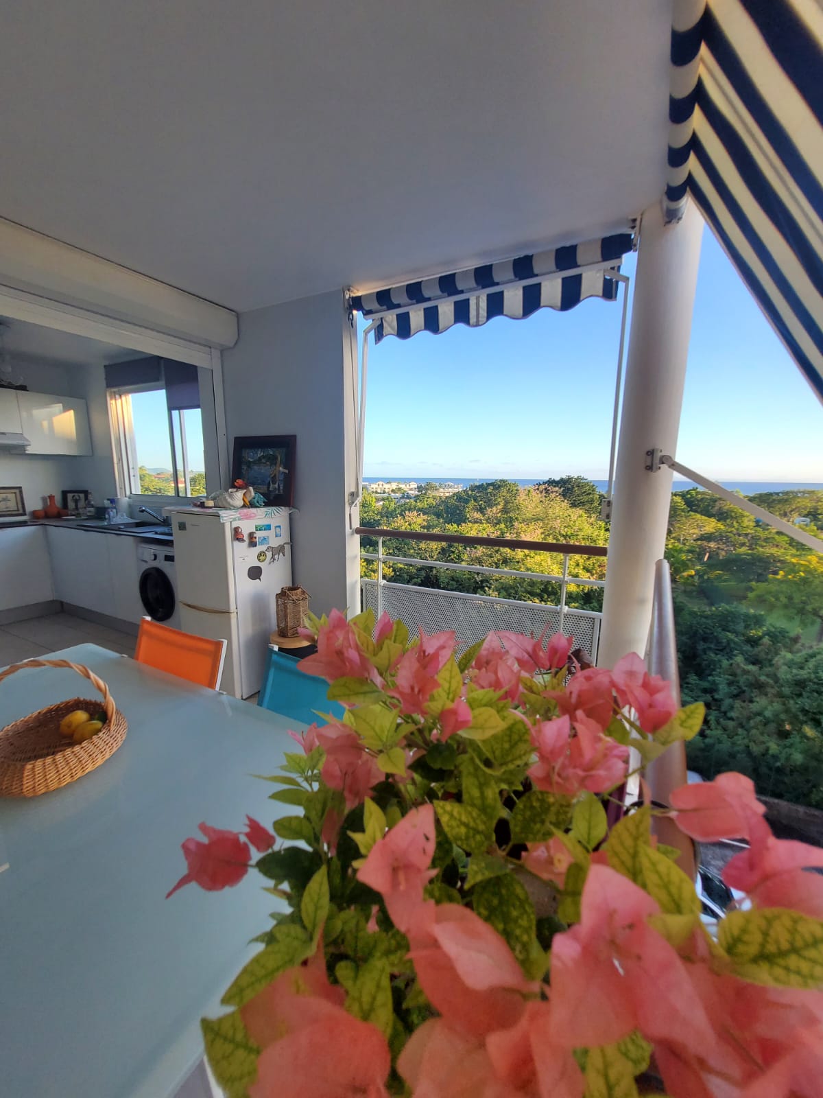 A bright dining area features a table with a view of the Caribbean Sea through large open doors. Bougainvillea flowers add a splash of color in the foreground, while lush greenery is visible outside, enhancing the connection with nature.