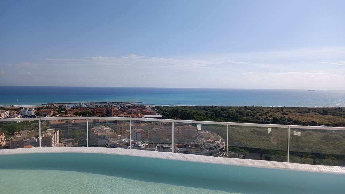 A panoramic view captures the serene expanse of the Mediterranean Sea, with gentle waves and a clear blue sky. A glass railing surrounds an infinity pool, enhancing the sense of openness. The coastal landscape below is visible, showcasing a mix of urban and natural elements.