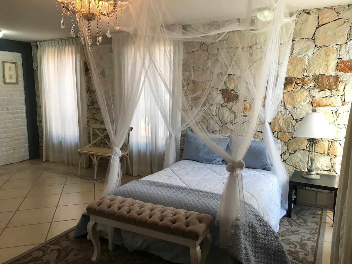 A spacious bedroom features a large bed draped with a soft canopy. The textured stone wall adds a natural element to the room. Light enters through sheer curtains, and a stylish chandelier hangs overhead. A bench sits at the foot of the bed, enhancing the room's charm.