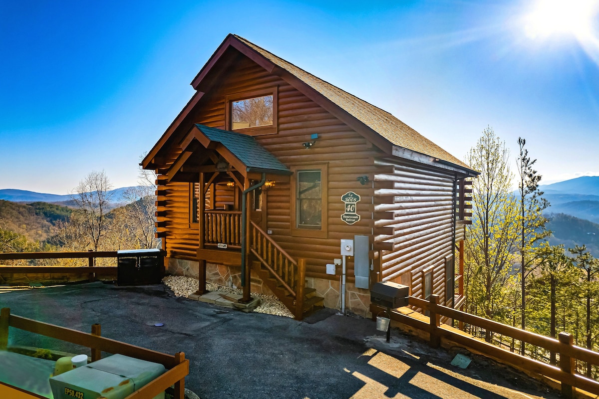 A modern rustic cabin exterior is showcased against a bright blue sky. Large wooden beams and logs define the charming structure, while a welcoming front porch leads to the entrance. Surrounding mountains are visible in the distance, complementing the serene setting.