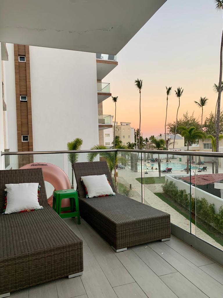 A private balcony is showcased with two lounge chairs overlooking the pool area. Palm trees can be seen in the distance against a soft, colorful sunset sky. The setting invites relaxation and enjoyment of the tranquil surroundings.