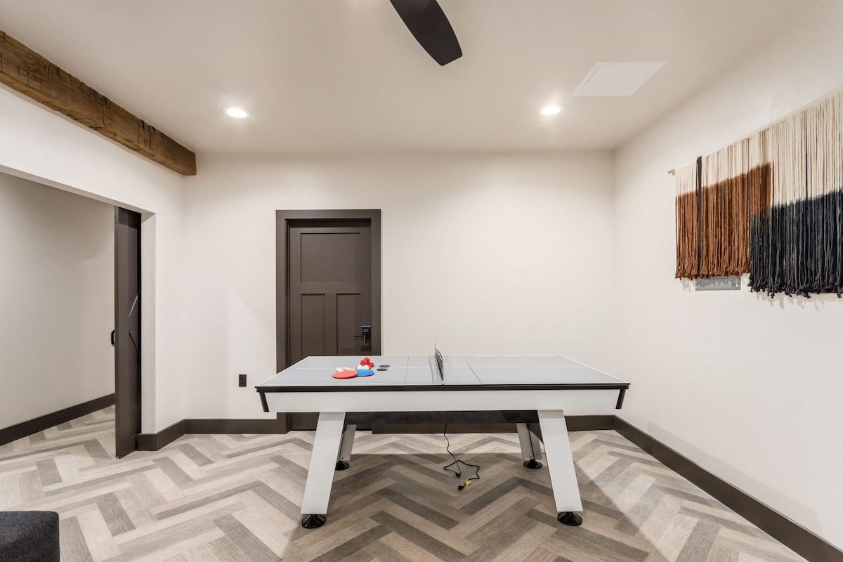 A game room is shown featuring a sleek ping pong table centered in the space. Light-colored walls highlight the contrasting dark door and ceiling fan. A textured wall hanging adds a warm touch to the neutral-toned decor and patterned flooring.