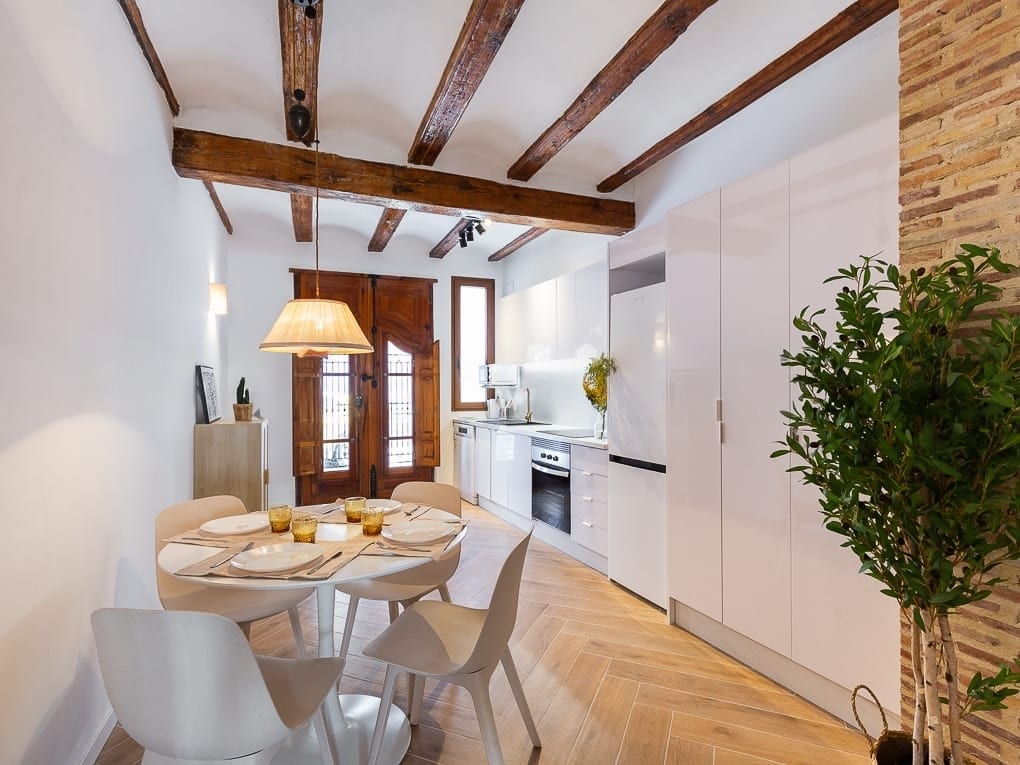 A modern kitchen area features a round dining table set for four, accompanied by sleek white chairs. Natural light filters through a wooden door, highlighting the warm tones of the tiled floor and the rustic wooden beams above.