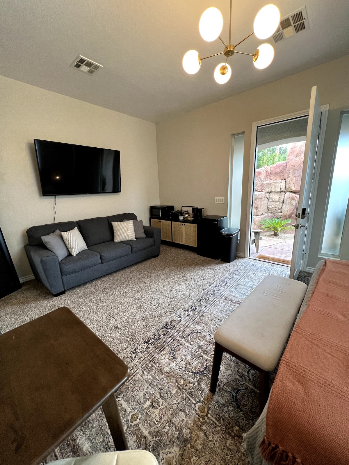 The living area features a comfortable couch with decorative pillows facing a wall-mounted television. A small table and a bench provide additional seating. Natural light is brought in through the glass doors, which lead outside, highlighting a soft area rug on the floor.