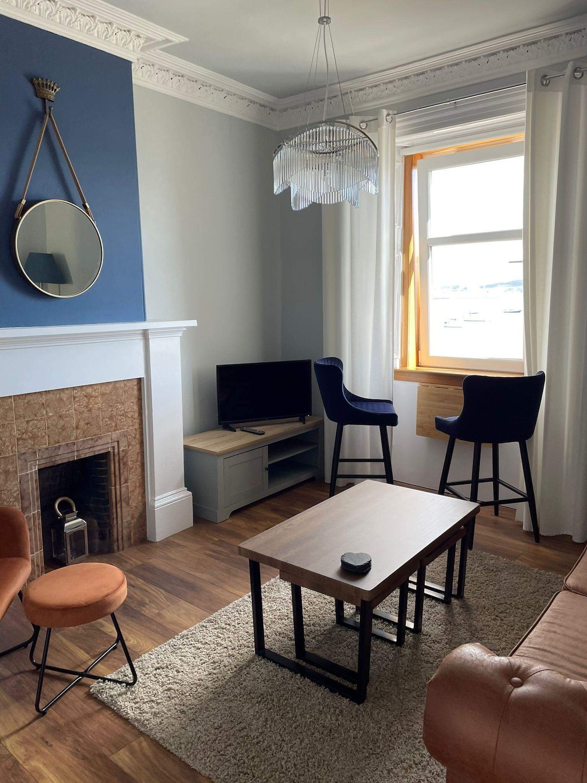 A cozy living area is highlighted by a stylish ceiling light fixture and a large window, allowing natural light to fill the space. Seating includes a sofa, two bar stools next to a small table, and a television on a console, complemented by a decorative fireplace.