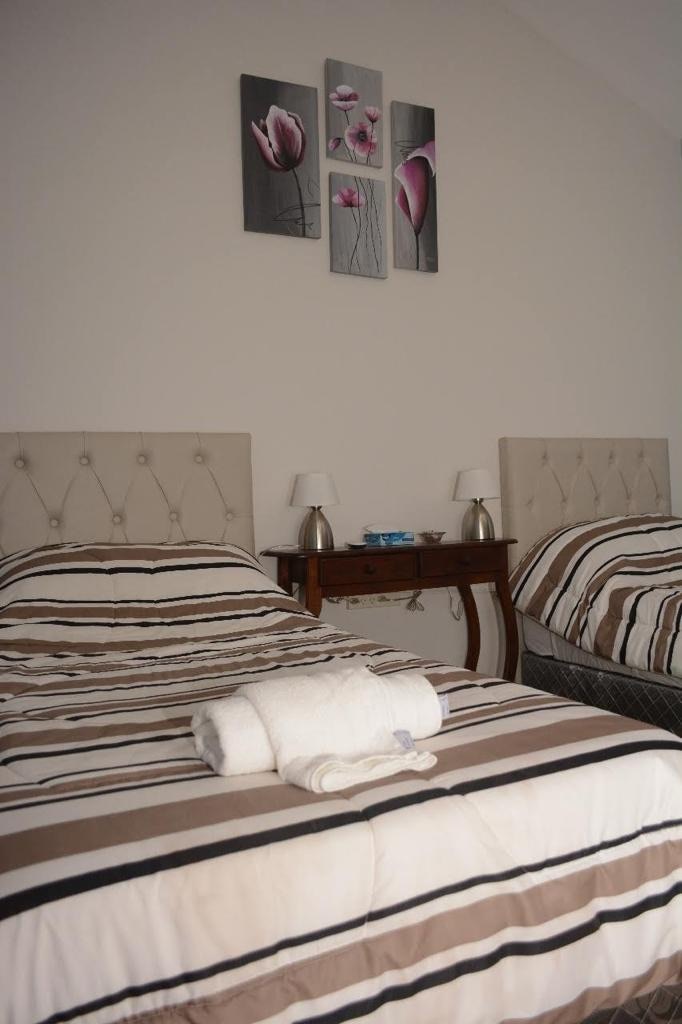The sleeping area features two neatly made beds with striped bedding in soft tones. Small bedside lamps are positioned on either side, complementing a decorative wall artwork of flowers. A wooden console table is placed between the beds, adding functionality to the space.