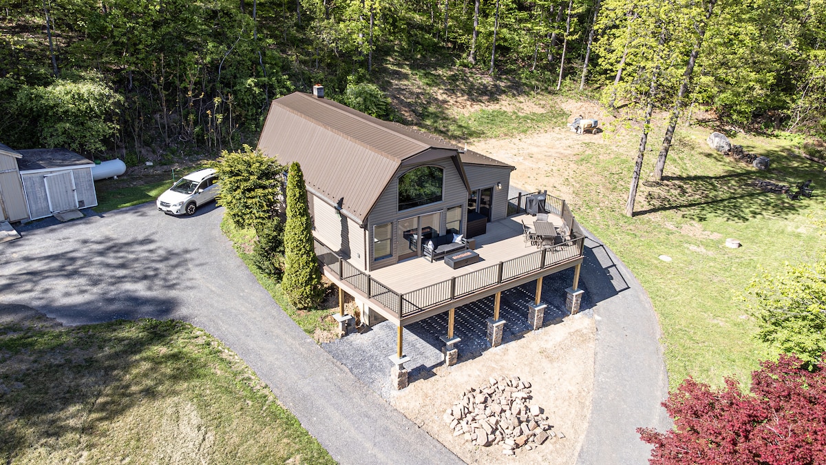 The contemporary cabin is situated on a spacious, natural setting, featuring a multi-level deck with outdoor seating. Nearby, a gravel path leads to the entry, while a vehicle is parked near the building. Lush greenery surrounds the property, creating a wooded backdrop.