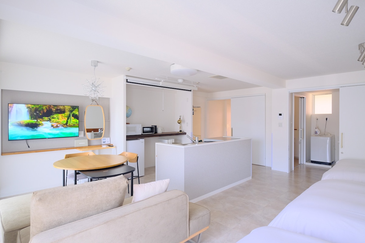An open concept living space features a comfortable lounge area with a light sofa, a dining table, and a large wall-mounted TV. A mini kitchen is visible, equipped with essential appliances, while a laundry area can be seen at the back through an open door.