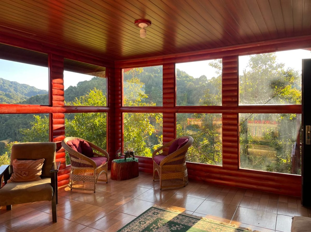 A sunlit sunroom is surrounded by large windows, offering views of the greenery outside. Two comfortable chairs with light upholstery and a small table occupy the space. Natural light fills the room, highlighting the wooden paneling and tile flooring.