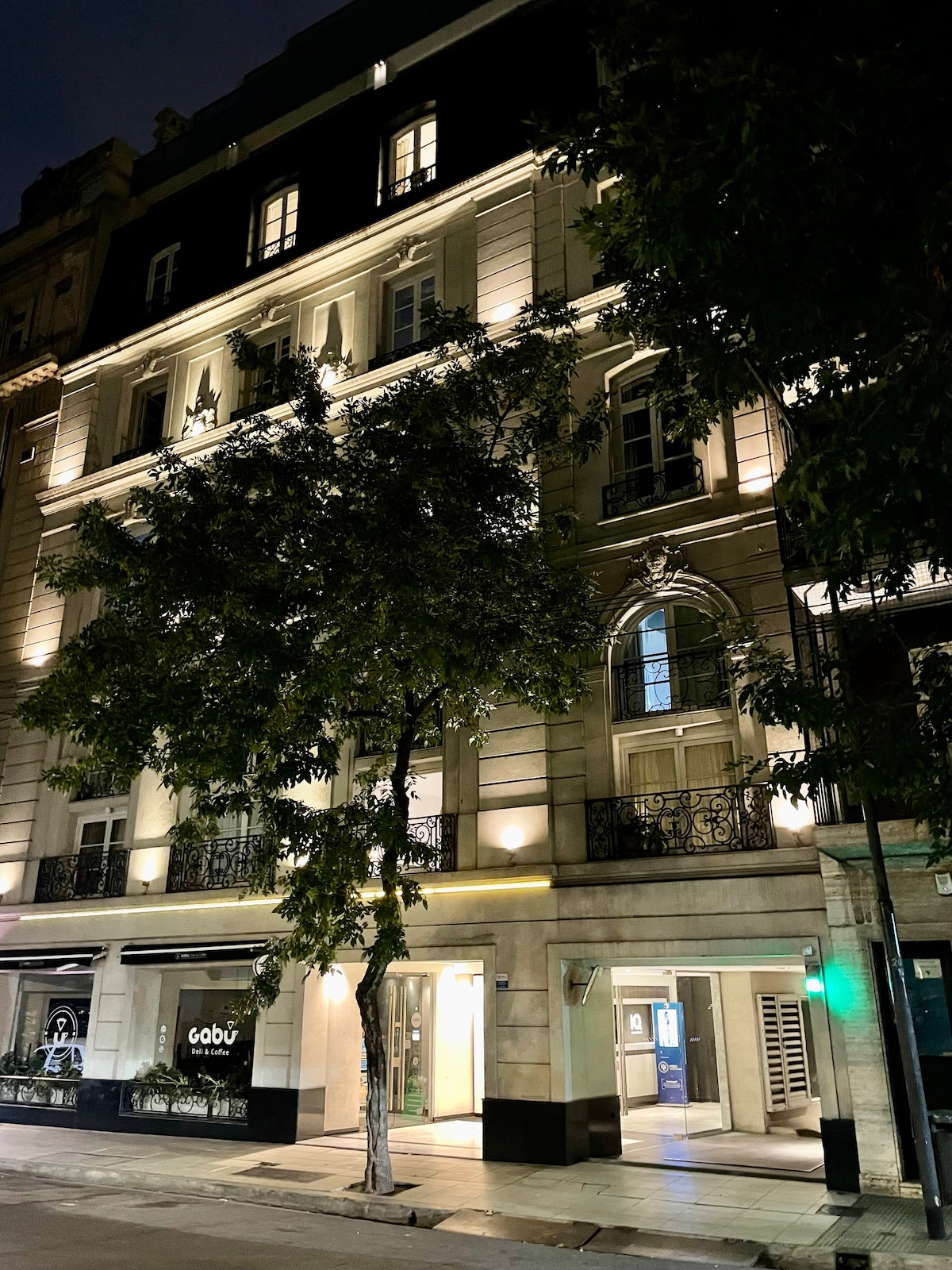 A historic building is illuminated at night, showcasing its elegant French architectural style. The façade features ornate balconies and large windows, framed by greenery on the sidewalk. Soft lighting highlights the intricate details of the structure, enhancing its presence in the upscale neighborhood.