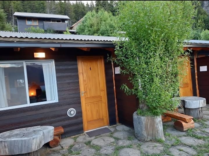 The entrance to a cozy accommodation is visible, featuring wooden doors and large planters filled with greenery. Stone pathways lead to rustic seating areas crafted from logs, creating an inviting outdoor space surrounded by trees.