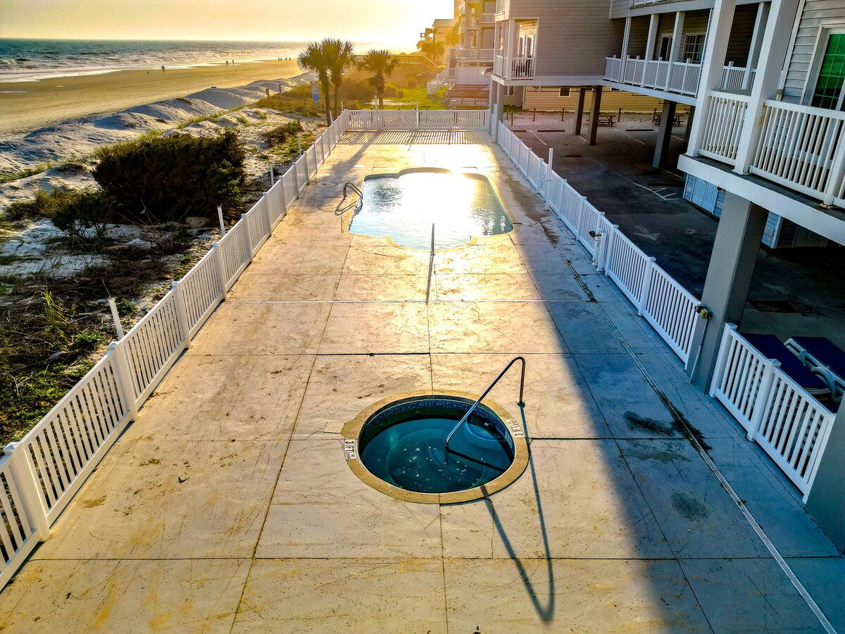 An outdoor pool area features a large swimming pool and a hot tub, both surrounded by a sunlit concrete patio. A white fence borders the area, and the ocean can be seen in the background, with gentle waves lapping at the shore.