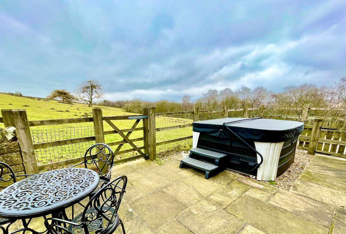A private patio area features a round table with four black chairs and a secluded hot tub covered for protection. A wooden fence encloses the space, providing views of the green fields and tree-lined countryside beyond, under a cloudy sky.