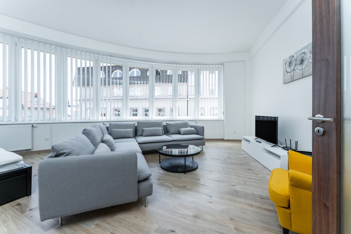 A light-filled living area features large windows with vertical blinds, providing an airy atmosphere. A modern gray sectional sofa is positioned around a sleek black coffee table. A television unit sits along one wall, and a vibrant yellow armchair adds a pop of color to the space.
