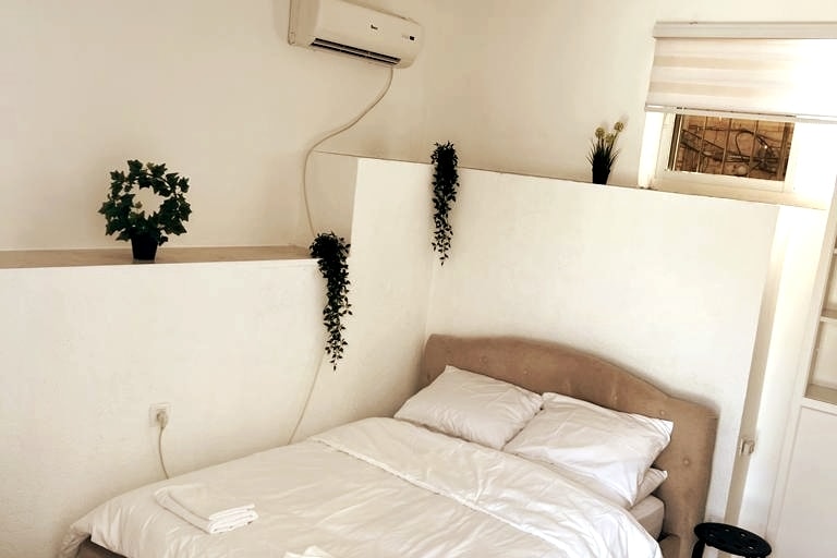 A bright sleeping area features a spacious bed with crisp white linens. Decorative plants are placed on the walls, adding a touch of greenery. Natural light filters through a window, while a mounted air conditioning unit ensures comfort.