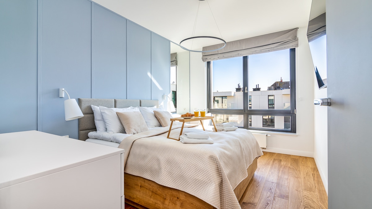 A serene bedroom features a spacious bed adorned with crisp linens and a light blanket. Natural light streams through large windows, highlighting the soft blue wall panels. A bedside table holds a tray with breakfast essentials, offering a welcoming touch to the space.