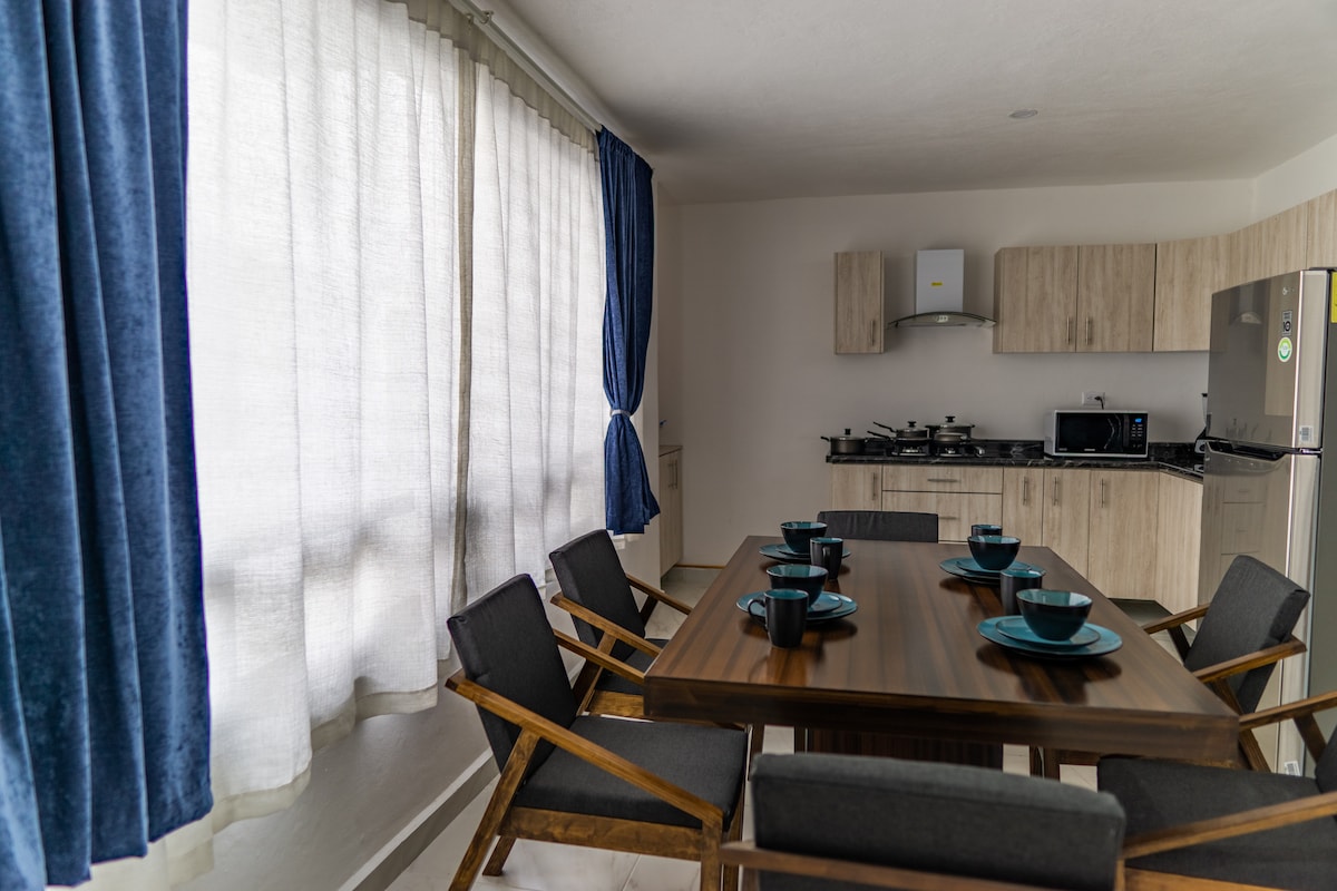 An inviting kitchen area is presented, featuring a wooden dining table surrounded by six chair-like seats. Natural light streams through floor-length curtains, illuminating the sleek cabinetry and modern appliances, including a refrigerator and stovetop. Dinnerware and utensils are neatly arranged at the table.