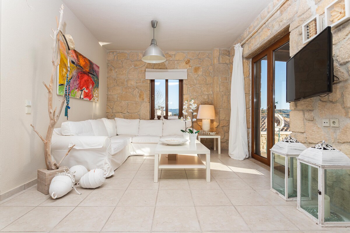 A light-filled living area features a white sectional sofa and a coffee table centered on a tiled floor. A decorative wall made of stone adds texture, while large windows offer natural light. A television mounted on the wall complements the modern decor.