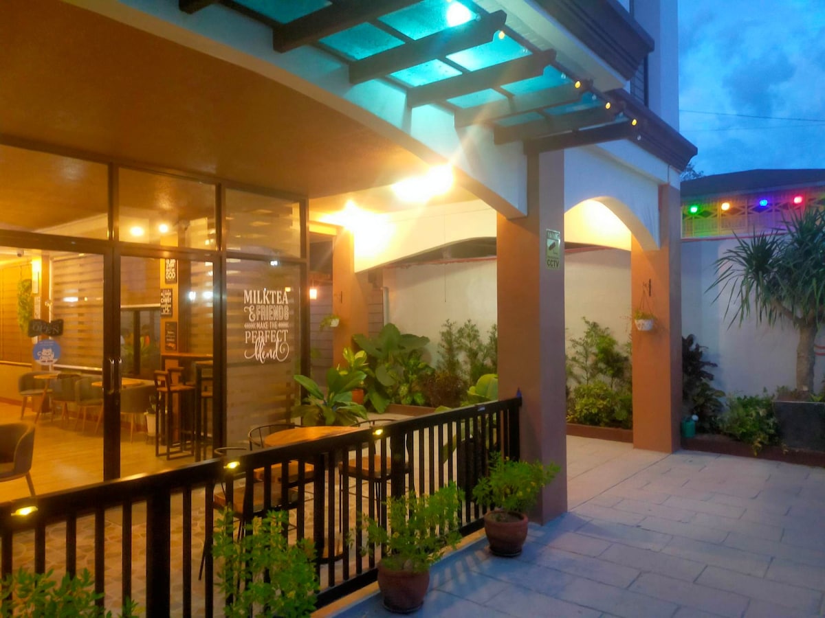 An inviting entrance features a covered outdoor seating area with modern furniture under soft lighting. Potted plants create a green border, while decorative accents on the walls enhance the welcoming vibe. The exterior is complemented by a colorful light display, visible against the evening sky.