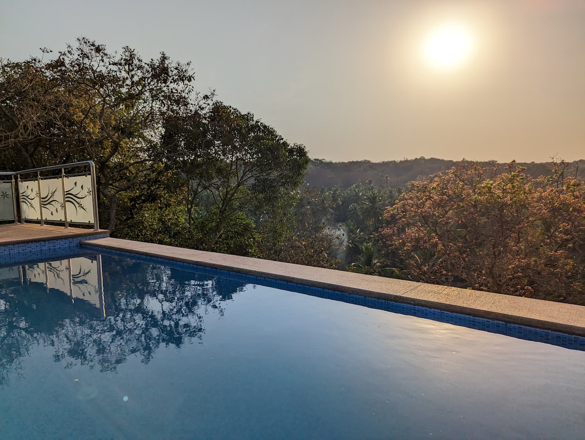 The infinity pool reflects the sun setting over lush greenery, creating a serene atmosphere. Surrounding trees add a natural backdrop, enhancing the peaceful ambiance. The calm water surface mirrors the colors of the sky, inviting relaxation and tranquility.