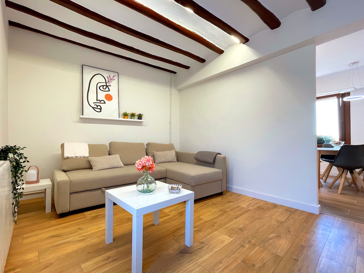 A bright living area features a spacious beige sofa and a minimalist white coffee table adorned with a small vase of pink flowers. Exposed wooden beams add character to the ceiling, while light enhances the warm wooden floor.