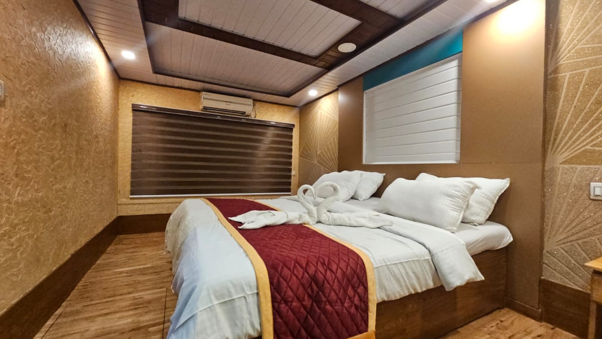 A comfortable sleeping area is highlighted by a spacious bed, featuring white linens and a maroon accent blanket. The room’s warm tones are complemented by soft lighting and a textured wall design, with shades drawn over a window allowing for controlled light.