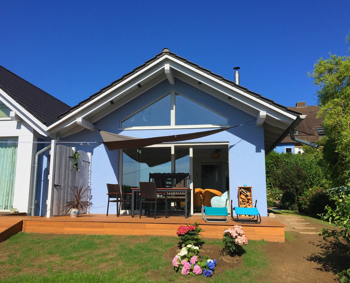 A light blue house with a modern design features large glass doors that showcase the interior. A spacious deck is furnished with a dining table and chairs, while a cozy seating area is visible. Colorful flowers, including pink and blue hydrangeas, are arranged in the garden.