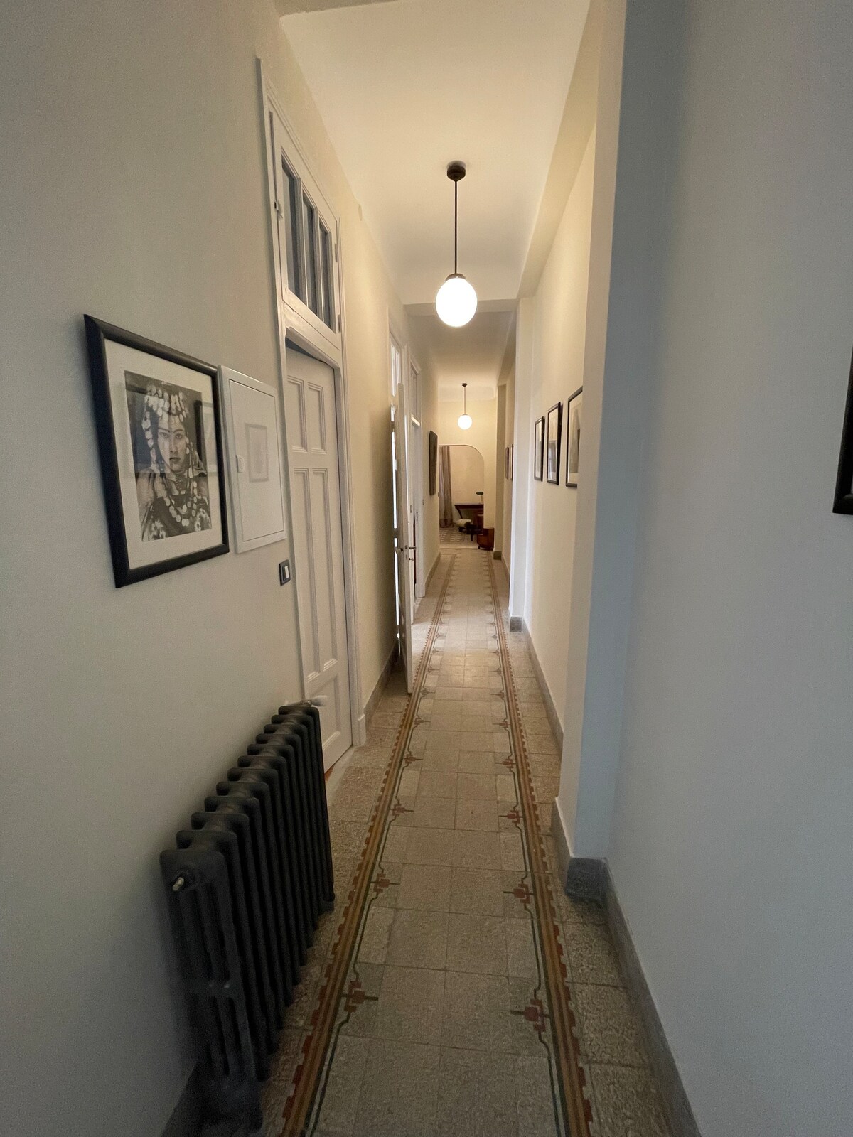 A long hallway features a central decorative tile pattern along the floor. Framed photographs line the walls, complementing the overhead pendant lighting. A radiator is situated on the left, and a glimpse of the living area is visible at the far end of the hall.