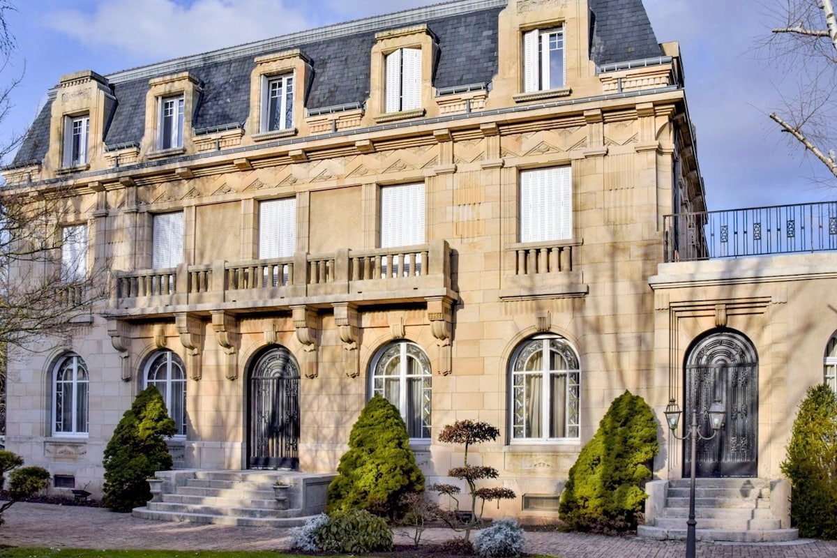 The exterior of a grand historical building showcases ornate architectural details and large windows. Lush green shrubs flank the entrance, while a set of stone steps leads to a pair of elegant black doors, enhancing the structure's stately presence.