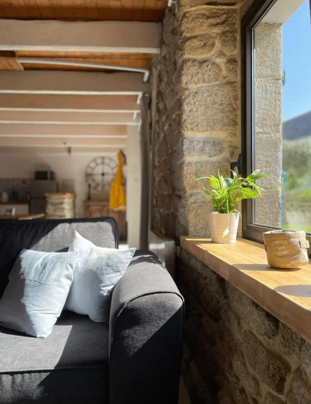Natural light illuminates the living space, highlighting a comfortable sofa adorned with soft cushions. A window ledge showcases a small potted plant and a decorative container, set against a backdrop of exposed stone walls and wooden beams.
